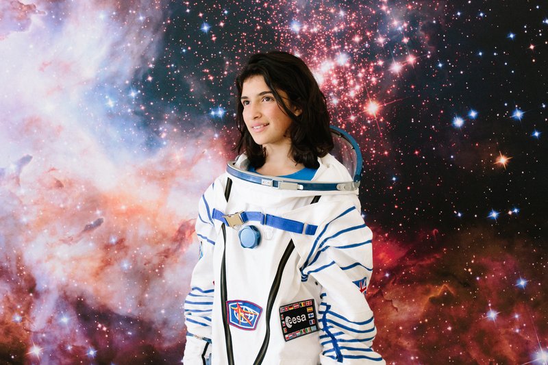 A teenage girl trying on a space flight suit, stood in front of a vibrant image of stars and giant gas clouds in space.