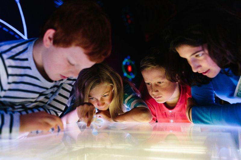 4 children gathered around an exhibit in a science centre, all looking down with fascinated looks at the interactive display in front of them