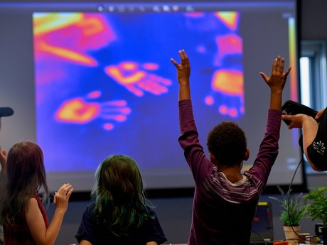 A group of children observe excitedly a projected image showing glowing hand prints left by heat on a surface viewed with an infrared camera.